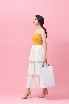 Full length of young woman holding shopping bags and smiling while walking isolated over pink background.