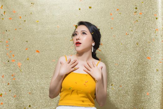 Portrait of a cheerful beautiful girl wearing dress standing under confetti rain and celebrating isolated over gold background