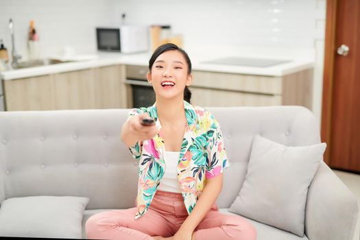 television, relax, home and happiness concept - smiling teenage girl sitting on couch with tv remote control at home