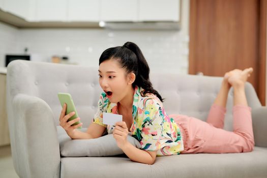 Image of optimistic excited young woman indoors at home using mobile phone holding credit card on sofa.