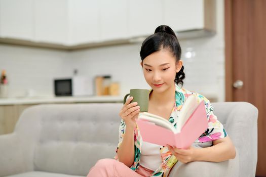 Young beautiful woman sitting on the sofa reading a book enjoying her tea in living room at home