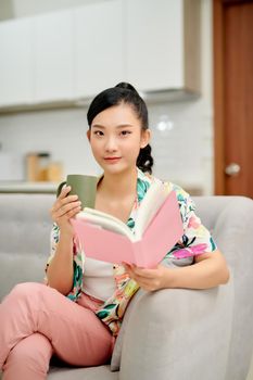 Young woman at home sitting on sofa relaxing in her living room reading book and drinking coffee.