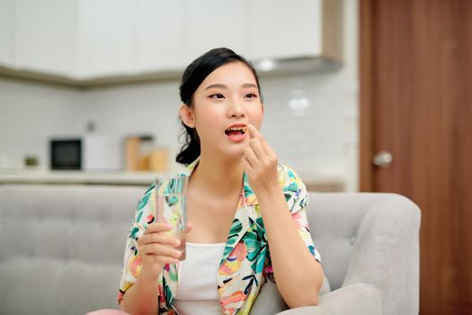 Asian woman holding pill and a glass of water sitting on sofa. 