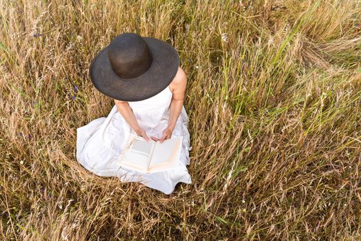 Romantic dreamy top view scene of unrecognizable woman sitting in a field of wheat and tall yellow grass wearing a dark wide-brimmed hat holding a book. Boho girl reading preferred romance story tale