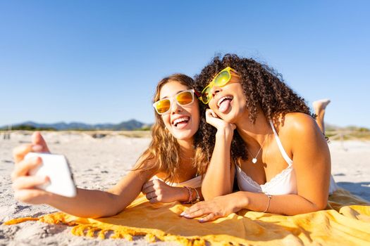Two beautiful girls lying on the beach in the summer having fun making faces with their tongues out while taking a selfie. Two multiracial lesbian girlfriends with funny sunglasses using smartphone