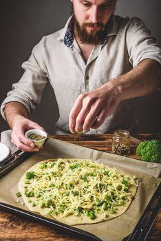 Bearded man pours spices on pizza with broccoli, pesto sauce and cheese