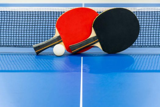 Black and red table tennis racket and a white ball on the blue ping pong table with a net, Two table tennis paddle is a sports competition equipment indoor activity and exercise for background concept