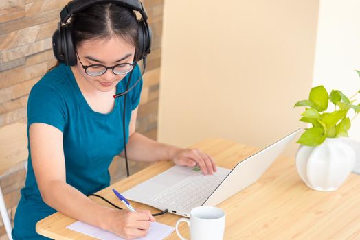 Asian teenage girl student with a headset is taking note learned online from school in a notebook using a laptop computer. Distance education from the class of the university by a video call from home