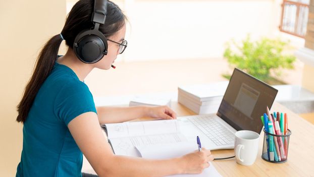 Asian teenage girl student with a headset looking at the laptop screen learning from school using a computer online. Distance education from the class of the university by a video call at a home on pc