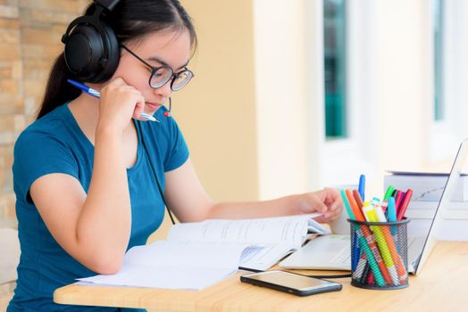 Asian woman student teenage girl with glasses headphones sitting looking serious reading a book worry using laptop computer on table learning online study. Education from a class of university at home