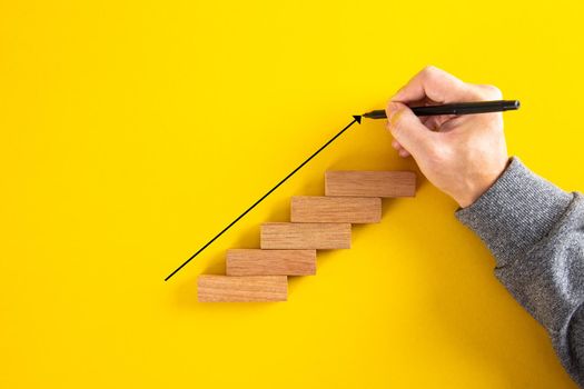 Man hand drawing an upward pointing arrow on top of growing graph made of wooden block. Over yellow background