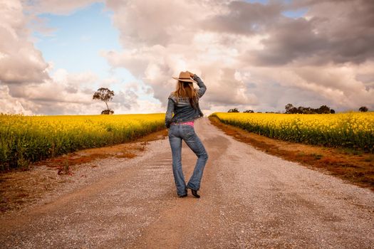 Aussie country girl wearing demin jeans and jacket standing in middle of a dirt road in farm fields of canola