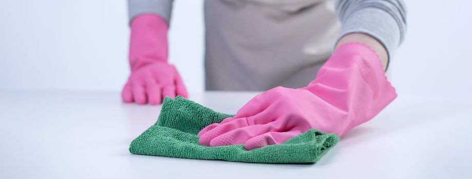 Young woman housekeeper in apron is cleaning, wiping down table surface with wet rag, spraying bottle cleaner to prevent disease spreading.
