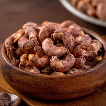 Cashew nuts with peel in a wooden bowl on wooden tray and table background, healthy raw food plate.