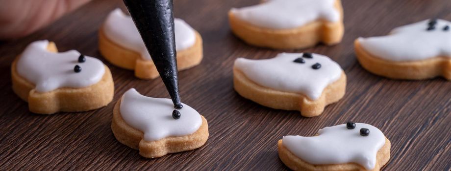 Close up of decorating cute Halloween ghost gingerbread cookies with frosting icing cream topping bag.