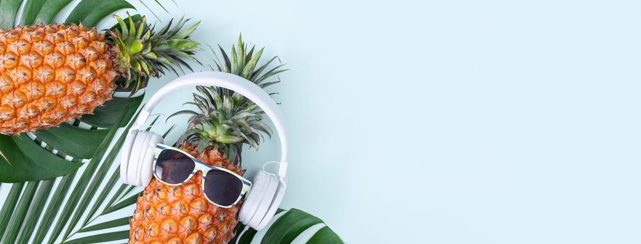 Funny pineapple wearing white headphone, concept of listening music, isolated on colored background with tropical palm leaves, top view, flat lay design.