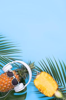 Funny pineapple wearing white headphone, concept of listening music, isolated on colored background with tropical palm leaves, top view, flat lay design.
