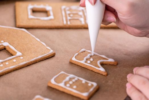 Young woman is decorating Christmas Gingerbread House cookies biscuit at home with frosting topping in icing bag, close up, lifestyle.