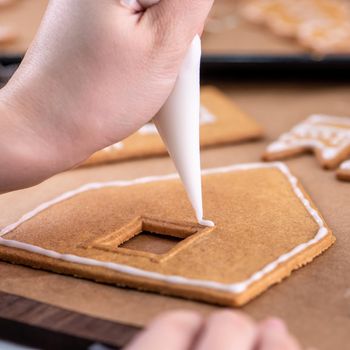 Young woman is decorating Christmas Gingerbread House cookies biscuit at home with frosting topping in icing bag, close up, lifestyle.