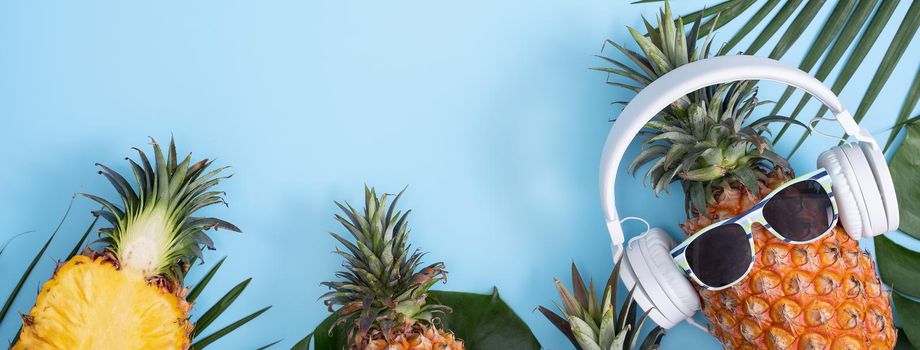 Funny pineapple wearing white headphone, concept of listening music, isolated on colored background with tropical palm leaves, top view, flat lay design.