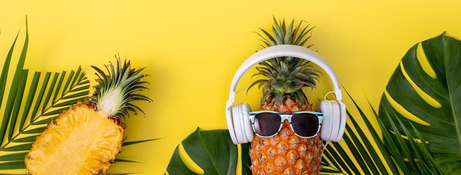 Funny pineapple wearing white headphone, listen music, isolated on yellow background with tropical palm leaves, top view, flat lay design concept.