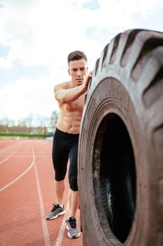 Young muscular man flipping a huge tire for training muscles at the tartan track on the stadium.