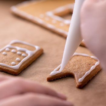 Young woman is decorating Christmas Gingerbread House cookies biscuit at home with frosting topping in icing bag, close up, lifestyle.