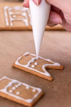 Young woman is decorating Christmas Gingerbread House cookies biscuit at home with frosting topping in icing bag, close up, lifestyle.