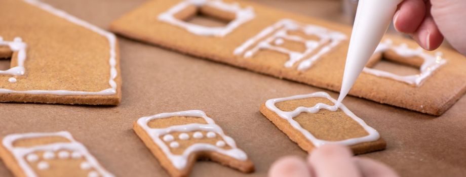 Young woman is decorating Christmas Gingerbread House cookies biscuit at home with frosting topping in icing bag, close up, lifestyle.