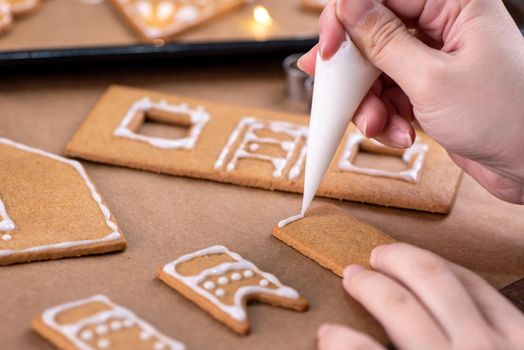 Young woman is decorating Christmas Gingerbread House cookies biscuit at home with frosting topping in icing bag, close up, lifestyle.