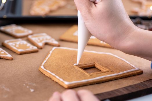 Young woman is decorating Christmas Gingerbread House cookies biscuit at home with frosting topping in icing bag, close up, lifestyle.