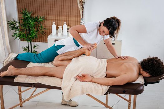 Handsome man lying down on massage table and getting healthy leg massage by young female therapist in the spa center.