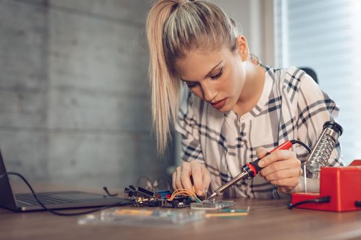 Young woman technician focused on the repair of electronic equipment by soldering iron.