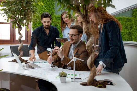 A team of a smart qualified skilled young engineers discussing about the project of alternative energy and one of them holding the tiny model of windmill turbine. 