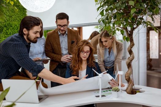 Young successful business colleagues analyzing project of alternative energy with miniatures of windmill turbines at the table, and  in a serious discussion, looking at paperwork. 