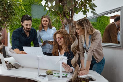 Young successful business colleagues analyzing project of alternative energy with miniatures of windmill turbines at the table, and  in a serious discussion, looking at paperwork. 