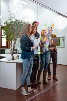 Creative professionals standing and discussing at the office behind glass wall with sticky notes and looking a post it note wall.