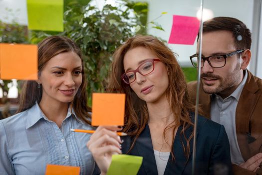 Creative professionals standing and discussing at the office behind glass wall with sticky notes and looking a post it note wall.
