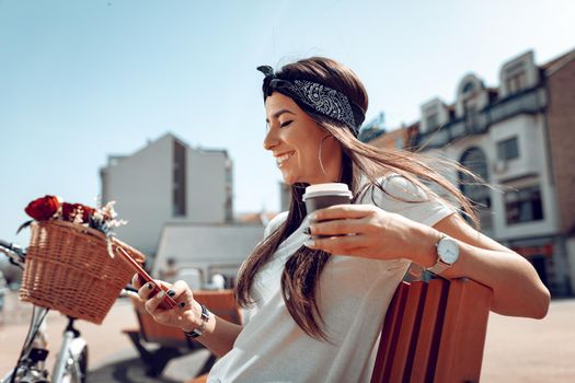 Smiling young woman with smartphone, taking coffee break and sits on a bench on a sunny day, beside the bike with flower basket.