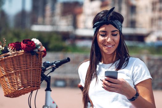 Smiling young woman texting on smart phone on the city street, on a sunny day, beside the bike with flower basket.