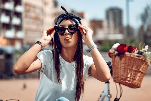 Pretty happy young woman is enjoying and having fun showing tongue, in a summer sunny day, beside the bike with flower basket.
