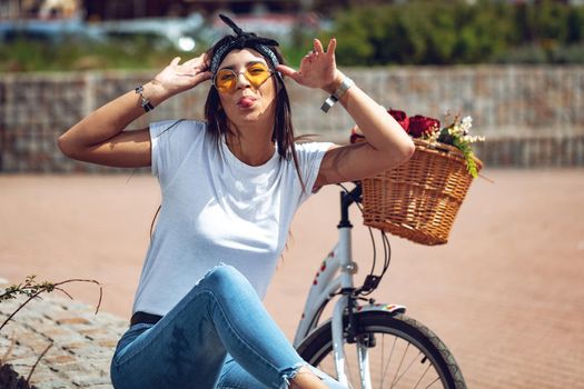 Pretty happy young woman is enyoing and having fun showing tongue, in a summer sunny day, beside the bike with flower basket.