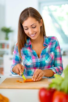 Beautiful young woman making healthy meal in the domestic kitchen. She is cutting carrot on the kitchen board.