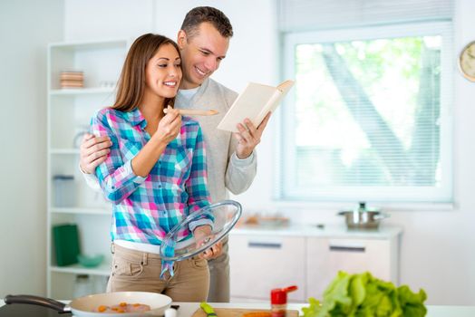 Beautiful young couple cooking healthy meal in the domestic kitchen. They are reading recipes from the cookbook.