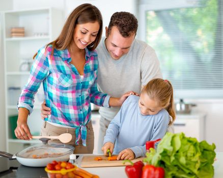Beautiful young family cooking healthy meal in the domestic kitchen. The little girl learns how to cut the carrot on the kitchen board and her parents supervise her.