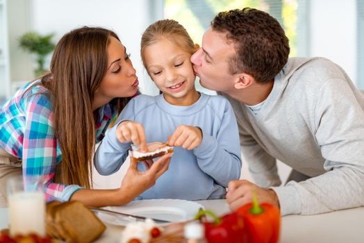 Beautiful young family preparing healthy meal for the breakfast in the domestic kitchen. The mother learns the little girl how to make the sandwich and father is kissing his daughter.