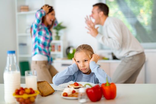Sad little girl having breakfast while parents quarreling in the kitchen. She is upset, holding head and looking away.      
