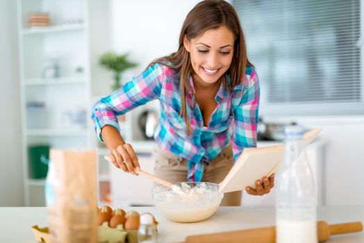 Beautiful young woman reading recipes from the cookbook and mixing dough in the dish for making the cookies for her family, in the domestic kitchen. 