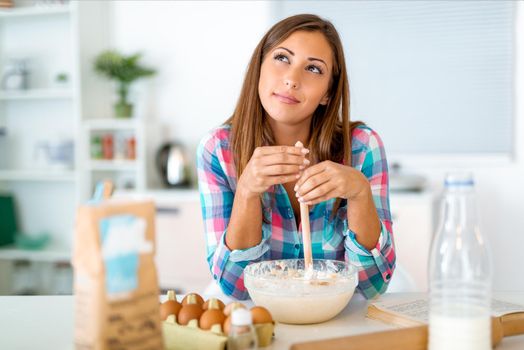 Beautiful young woman thinking what of components to put in dish with dough for making the cookies for her family, in the domestic kitchen. 