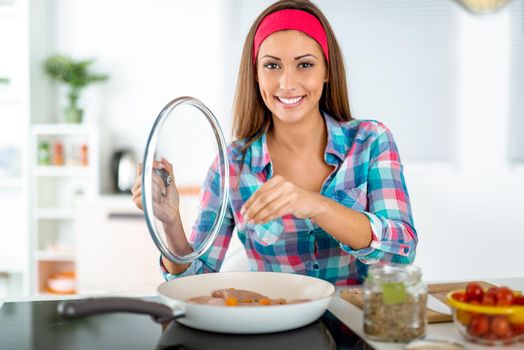 Beautiful young woman making healthy meal in the domestic kitchen. She is putting spices on chicken meat in frying pan. Looking at camera.